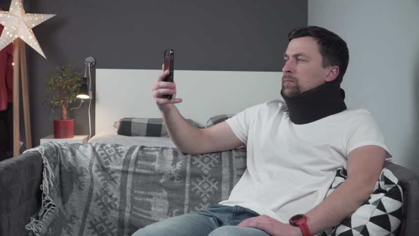Young man sitting at home on the couch talking on video calls about how he injured his neck and now needs rehabilitation and to wear a neck support collar. Cervical neck brace. Spinal trauma neck.  | Shutterstock HD Video #1109782527