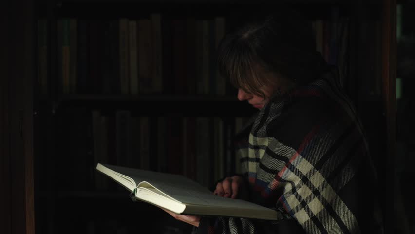 A woman is reading a book, in the background are shelves and many books. A homeschooler studies literature at home. A smart girl holds a book in her hands. Night atmosphere, close-up in retro style.