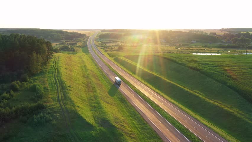 Long haul semi truck driving on intercity highway in rural region at sunset sun shines. Agricultural green crop fields and hills in background. Stage of delivery and logistics goods, aerial motion Royalty-Free Stock Footage #1109791683