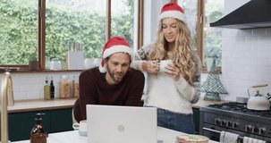 Diverse couple wearing santa hats using laptop for christmas video call, in slow motion. Relationship, togetherness, communication, celebration, christmas, lifestyle and domestic life, unaltered.