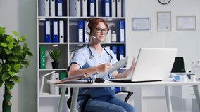 laboratory online, a young female medical worker in glasses communicates using a microphone and headphones via video communication on laptop while sitting in a hospital office