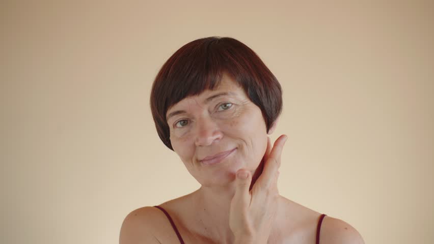 Happy smiling mature woman 50s skin care of the neck, neckline. Portrait of a beautiful middle-aged woman looking at camera, applying lifting anti-wrinkle cream moisturizing dry skin. Beige background