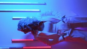 Vertical video. Young beautiful smiling hipster woman in street style fashion concept. Hot model wearing white suit and sunglasses. Female posing in blue neon light in studio interior