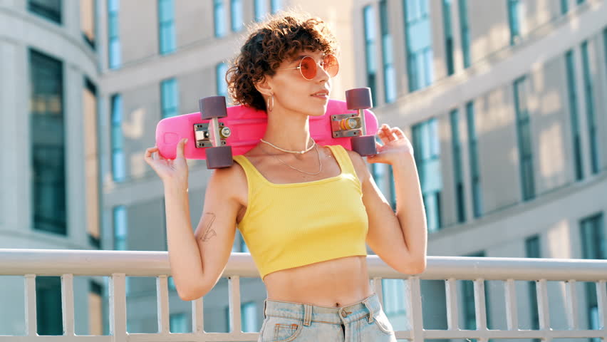 Young beautiful smiling hipster woman in trendy summer clothes. Carefree woman with curls hairstyle, posing in the street. Positive model outdoors. Cheerful and happy. Holds penny skateboard