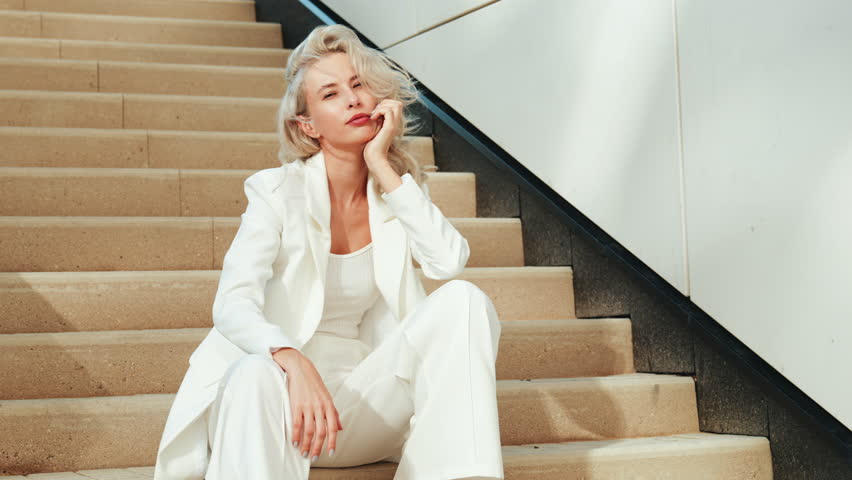 Young beautiful blond woman wearing nice trendy white suit jacket. Smiling model posing in the street at sunny day. Fashionable female outdoors. Cheerful and happy. sits at stairs