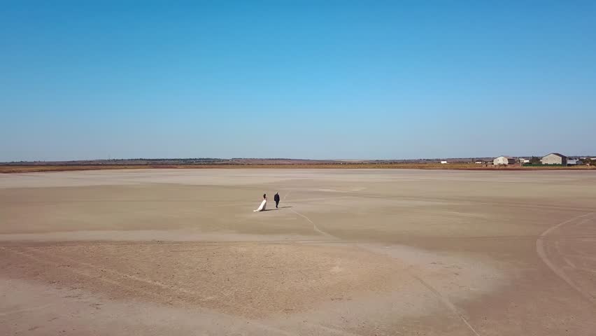 Aerial of newlyweds going at sandy desert landscape of dry lake. Bride in wedding dress, groom in suit walking at photoshoot. Just married man, woman enjoy wedding day. Drone video, global warming.