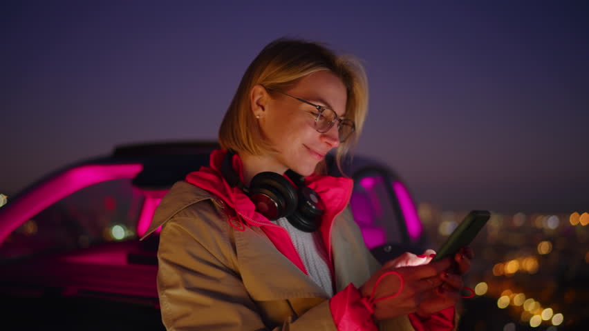 Caucasian female traveler sitting in car interior at night time and using smartphone application. Young cheerful woman sending text messages to friends in online chat, while browsing social media page