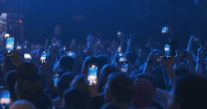 Fans crowd filming musical group performances on phones. Blue phone screens in people hands record live event entertainment on bright orange light background. Happy audience enjoy show in nightclub