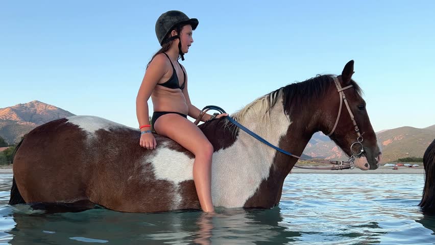 Young red-haired girl in bikini have fun bathing and bareback riding horse in sea water in summer season
