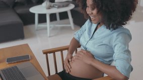 a joyful pregnant African young adult female at home. She is engaged in a video call via a laptop, sharing the ultrasound picture of baby in the womb with husband and family.