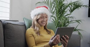 Happy biracial woman wearing santa hat using tablet for christmas video call, in slow motion. Communication, celebration, christmas, lifestyle and domestic life, unaltered.