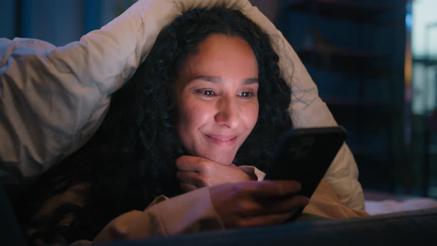 Happy woman gadget addict social media addicted Hispanic smiling girl Latino Indian lady Arabian female suffering insomnia at night home relaxing under blanket cover with duvet browsing mobile phone Royalty-Free Stock Footage #1109825057