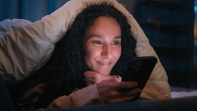 Happy woman gadget addict social media addicted Hispanic smiling girl Latino Indian lady Arabian female suffering insomnia at night home relaxing under blanket cover with duvet browsing mobile phone - Powered by Shutterstock - Get 15% off with code: PIKWIZARD15