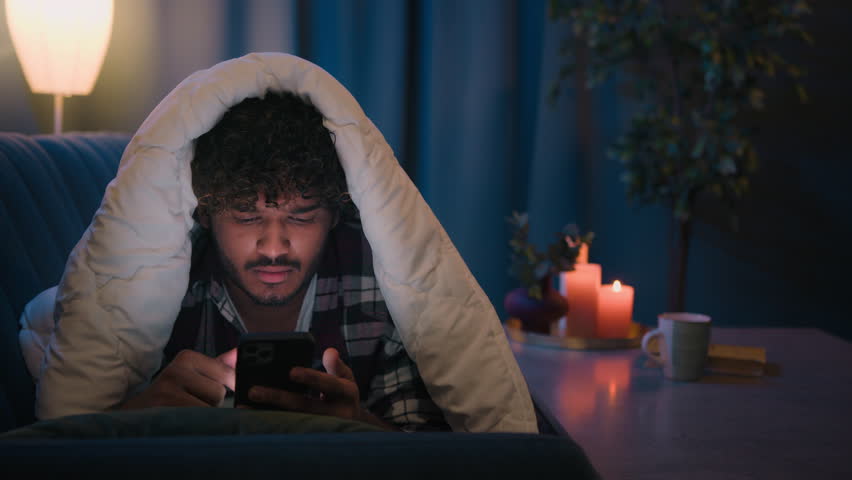 Night at home Hispanic guy gadget internet social media addict tired sleepy man reading news in social media shocked confused puzzled exhausted Arabian Indian male under blanket with mobile phone app Royalty-Free Stock Footage #1109825061