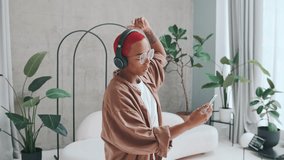 Young charismatic pretty African American woman blogger in headphones dances and takes selfie video on mobile phone enjoying online communication with subscribers located inside stylish house.