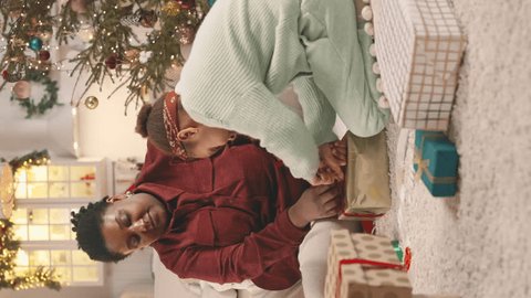 Vertical shot of playful African American little girl helping mom to tie bow on Christmas present with red ribbon, sitting together on floor in cozy decorated apartment ஸ்டாக் வீடியோ