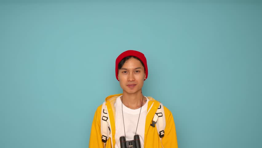 Portrait of a young Asian man in a yellow jacket and red cap pointing with his finger on a blue background