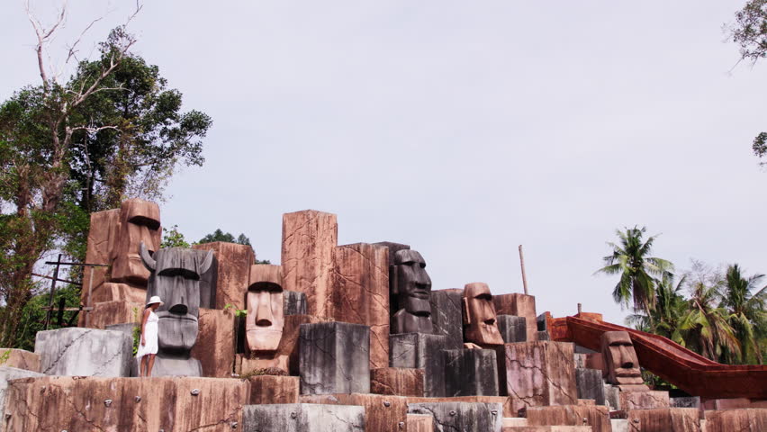 A woman gracefully strolls atop stone pedestals amidst stoic head statues in an abandoned hotel pool during daytime in Ko Chang, Thailand