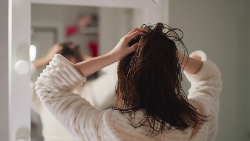 Female in robe twirls wet flying hair in lighted bathroom slow motion. Woman moves hands adjusting hair in bathroom with blurry reflection in mirror