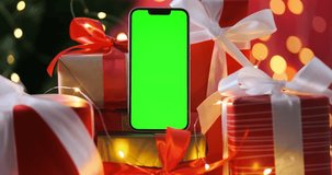 Smartphone screen with chroma key in Christmas gifts on golden-red glowing garland backdrop, close up. Phone green screen on Christmas tree background. Christmas spirit. Holidays celebration. 4k video
