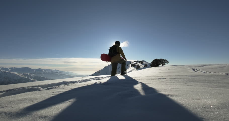 Snowboarder walks along mountain snowdrifts. Backcountry freerider snowboarder athlete sportsman walks against of clear sunny sky to peak of mountain. Climbing mountain for descent on snowboard