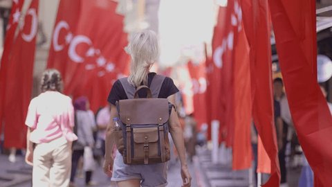 Back view Happy tourist blonde woman with backpack walking on old street turkish red flags. Tourist traveler discovers interesting places and popular attractions and walks around old city of Istanbul - Βίντεο στοκ