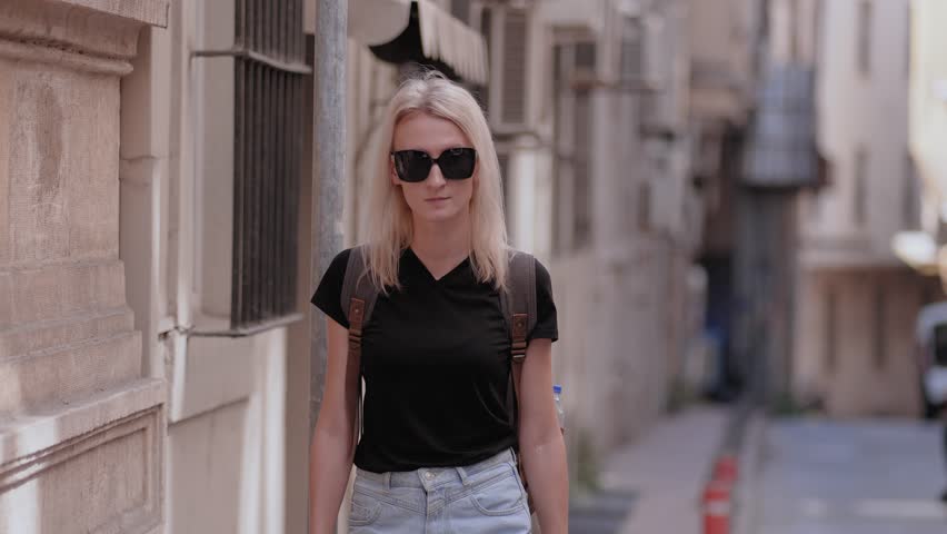 Front view of blonde tourist woman wearing dark sunglasses with backpack. She walks, walks down old empty street and enjoys journey and new place, sightseeing. Green spaces and trees along road.