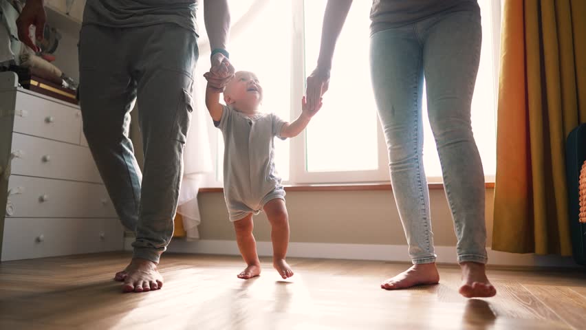 First steps. Family day. toddler takes first steps at home across field. parents hold hands of baby learn to walk. Happy baby takes first steps on floor of house.toddler dream to learn the first steps Royalty-Free Stock Footage #1109845471