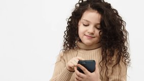 A small, curly-haired girl uses a phone on a light background. The concept of modern technology and internet communication.