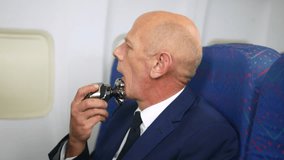 A mature business man using an electric razon to shave his face on a plane. The senior male is sitting in the passenger cabin of an airliner whist grooming - Powered by Shutterstock - Get 15% off with code: PIKWIZARD15