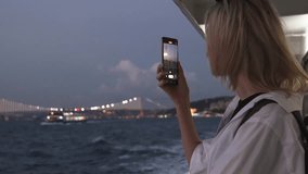 Young beautiful businesswoman blonde woman in sunglasses on ferry in Istanbul plfving on Bosphorus. Girl in white stylish shirt riding carbel or boat and taking pictures with smartphone. Sightseeing