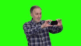 Happy mature man taking photos on his smartphone and swiping them. Isolated on green.