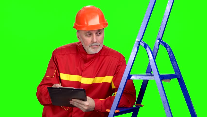 Mature caucasian contruction worker in red uniform writes down something on clipboard and talking. Isolated on green.