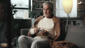 Excited mature gray haired man gamer sitting on a couch and playing in video games on a console at home Happy senior player male controlling joystick for online competition indoors