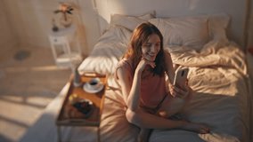 Morning woman video calling in golden sunny bedroom. Chill girl resting hotel room talk mobile phone at cozy home. Smiling relaxed teenager videocalling use smartphone in bed. Weekend routine concept