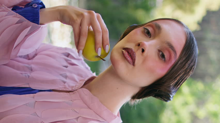 Sensual fantasy model posing at forest lake holding green apple close up. Gorgeous fairy girl sitting at mesmerizing nature place. Magical woman portrait closing eyes enjoying fairytale vertical view.