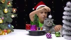 On a black background with flashing lights, a child in a Santa Claus hat and a cat play with New Year's decorations