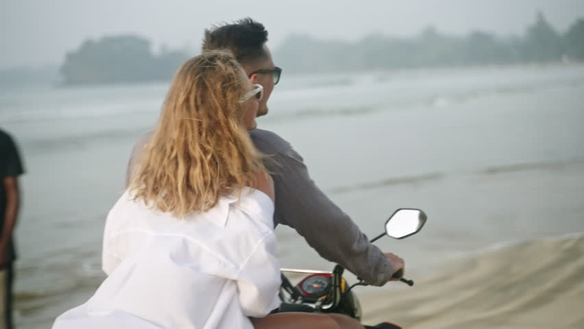 Happy couple riding motor bike on seaside at sunrise. Man, woman driving motorcycle on ocean coast. Pretty lady in bikini, shirt, sunglasses looks back to camera, laughs, smiles. Wind blows hair.