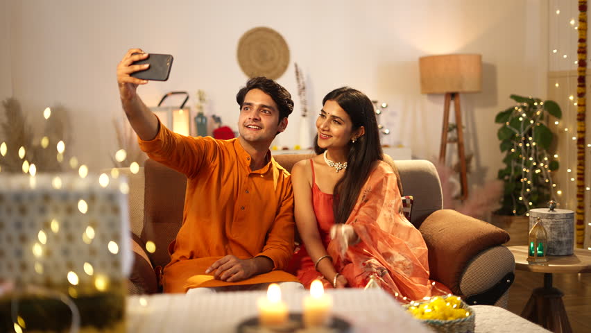 Indian beautiful couple click a selfie picture while sitting on the sofa.wife is doing flying kiss,well decorated home,hindu festival concept.