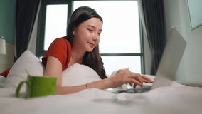 Attractive young Asian freelancer woman with a laptop on the bed in a condo near window. Travel and remote work at home concept with free schedule.