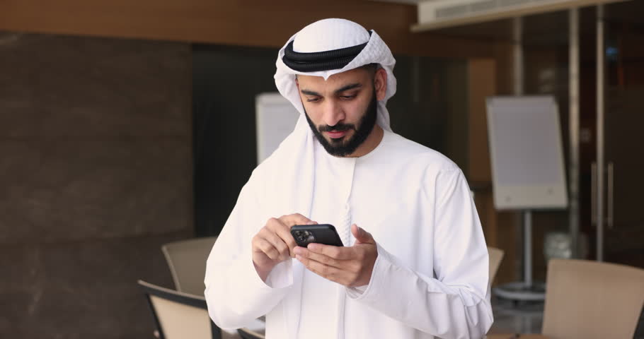 Millennial Muslim Indian office employee using smartphone. Businessman in kandura hold modern gadget, solve business, check e-mail, answer to client by sms, using new mobile business app at workplace Royalty-Free Stock Footage #1109893511