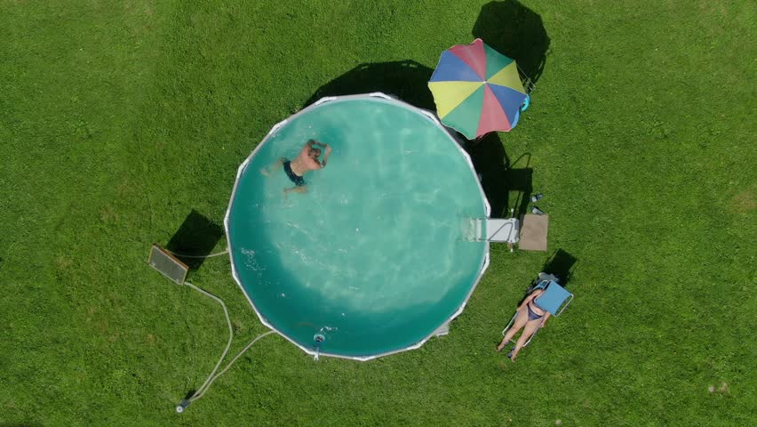 Aerial view of the man swimming in the pool and woman taking a sunbath on the backyard on the green lawn