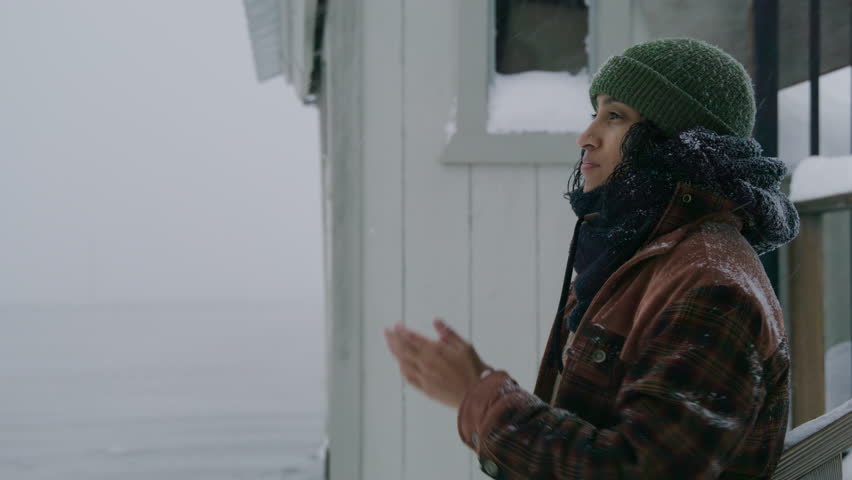 Medium profile shot of thoughtful Indian woman in plaid jacket, green hat standing near house at lake during blizzard, robbing hands, getting warm. High quality 4k footage Royalty-Free Stock Footage #1109894653