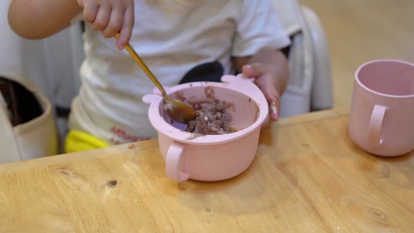 Acion of a baby is using little spoon to eating a scoop of chocolate icecream which is in pink plastic bowl and placed on wooden table. Baby lifetyle and learning practice, 4k footage.