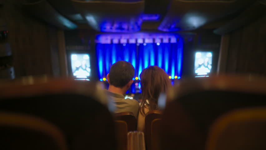 Young couple sits in chairs in half-empty concert hall before show starts, stage illuminated with blue lights. They using smartphone, taking selfies or checking social networks Royalty-Free Stock Footage #1109907279