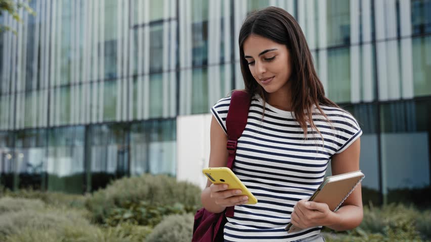 Happy hispanic student girl using yellow cell phone outdoors at University campus. Smiling young woman having fun with online app  Royalty-Free Stock Footage #1109912619