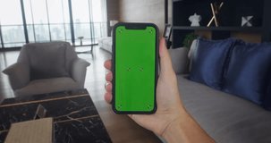 Male hand holding mobile phone with vertical green screen on living room. Male using mobile phone with green screen for copy space. Chromakey mock up. Browsing Internet, Watching Video Content, Blogs.