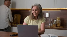 Mature 50s business woman wife online working with computer distance learning using laptop and mobile phone devices while senior man husband washing dishes in kitchen middle aged family couple at home - Powered by Shutterstock - Get 15% off with code: PIKWIZARD15