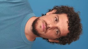 Vertical video, Close-up of guy with curly hair dressed in blue t-shirt, cheerful guy speaking sharing emotions on camera, video blog, live stream, virtual communication on blue background 