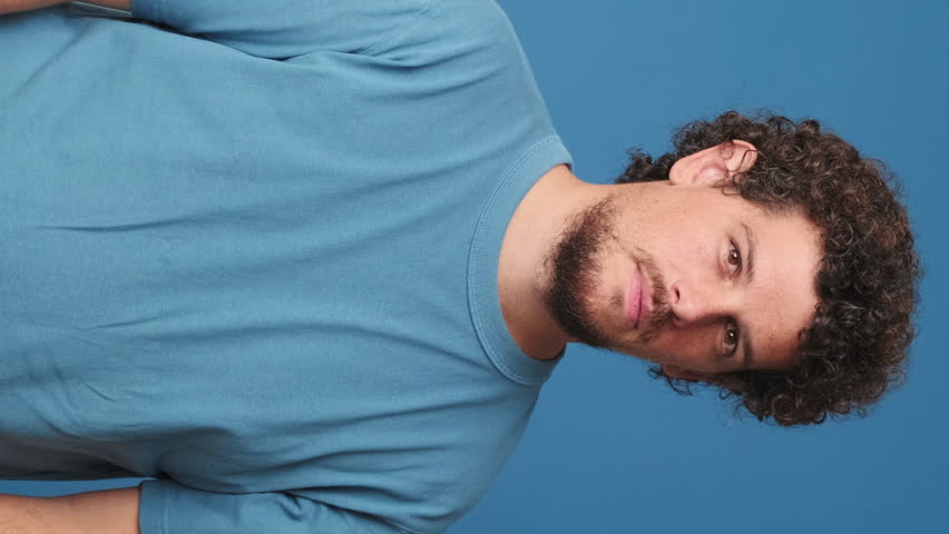 Vertical video, Happy guy with curly hair dressed in blue t-shirt looking at camera and showing sign of shape heart isolated on blue background in studio